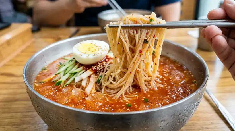 Macro shot of wheat noodles lifted by chopsticks from icy Milmyeon broth with egg and cucumber in steel bowl
