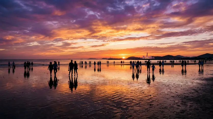 Dramatic purple, orange, and gold sky reflected on wet sand with silhouettes of people and fountain
