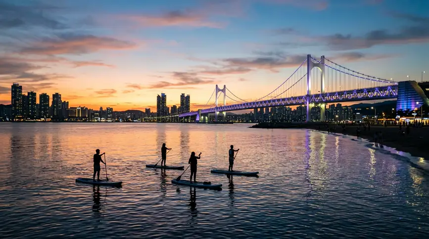 Silhouettes of people on paddleboards with Gwangan Bridge lighting up in background at sunset