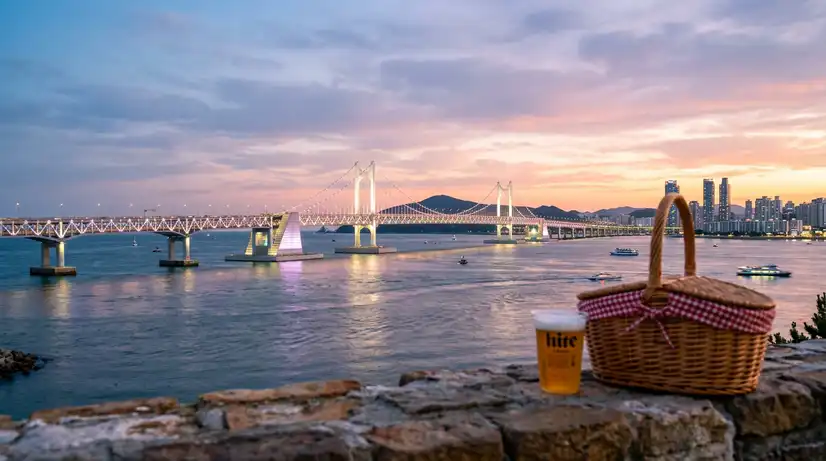 Sunset view of Gwangan Bridge with twinkling lights and blurred foreground showing picnic basket and beer on ledge
