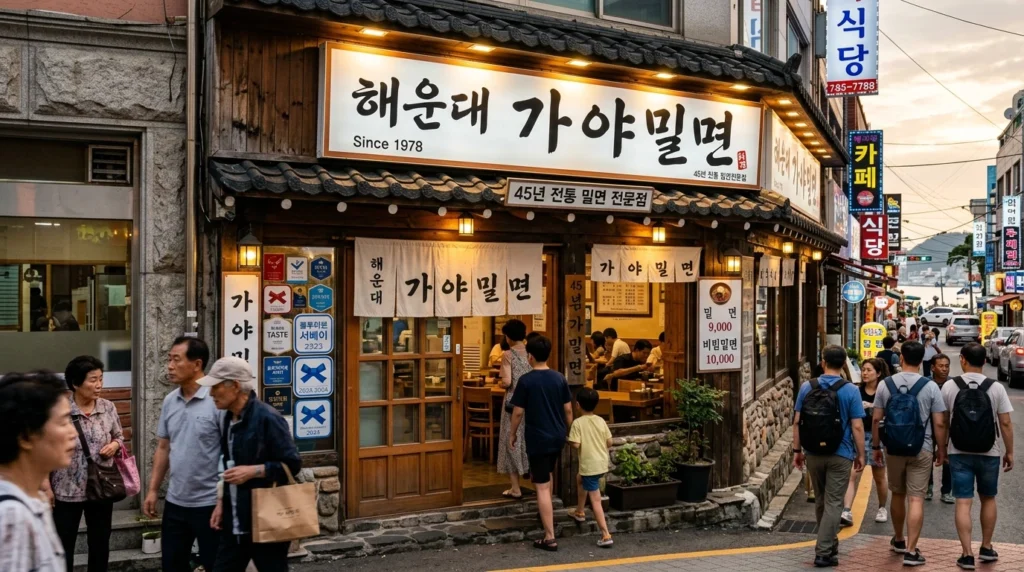 Professional documentary-style photography showing Haeundae Gaya Milmyeon restaurant storefront near beach area with traditional Korean restaurant facade and prominent Korean signage, busy street scene with tourists and locals, Blue Ribbon Survey award visible, cultural heritage atmosphere with warm inviting lighting and established authentic restaurant appearance 