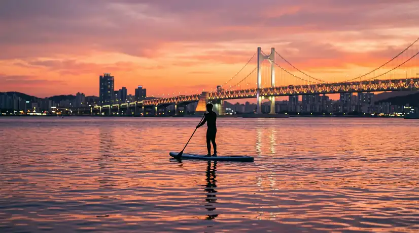 Peaceful sunset with person on paddleboard silhouette, orange-pink sky, illuminated Gwangandaegyo Bridge background